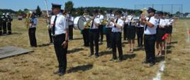 Zawody sportowo-pożarnicze w Jeziorach. Plac ćwiczeń. Na tle lasu stoi orkiestra dęta, na czele z kapelmistrzem. Muzycy wykonują utwór. Na placu widoczne sa przeszkody i chorągiewkikoloru czerwonego. 