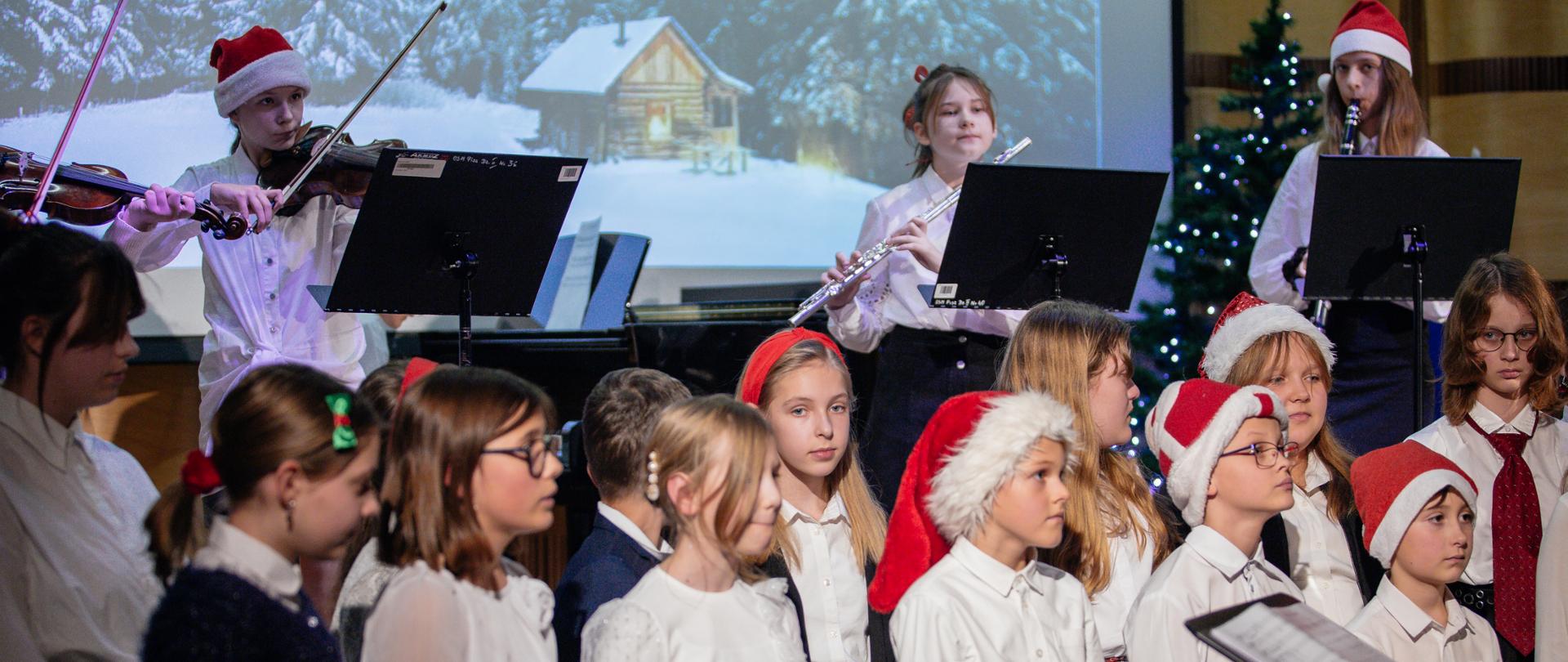 Zdjęcie chóry dzieci przebranego świątecznie, w tle zespół instrumentalny. 