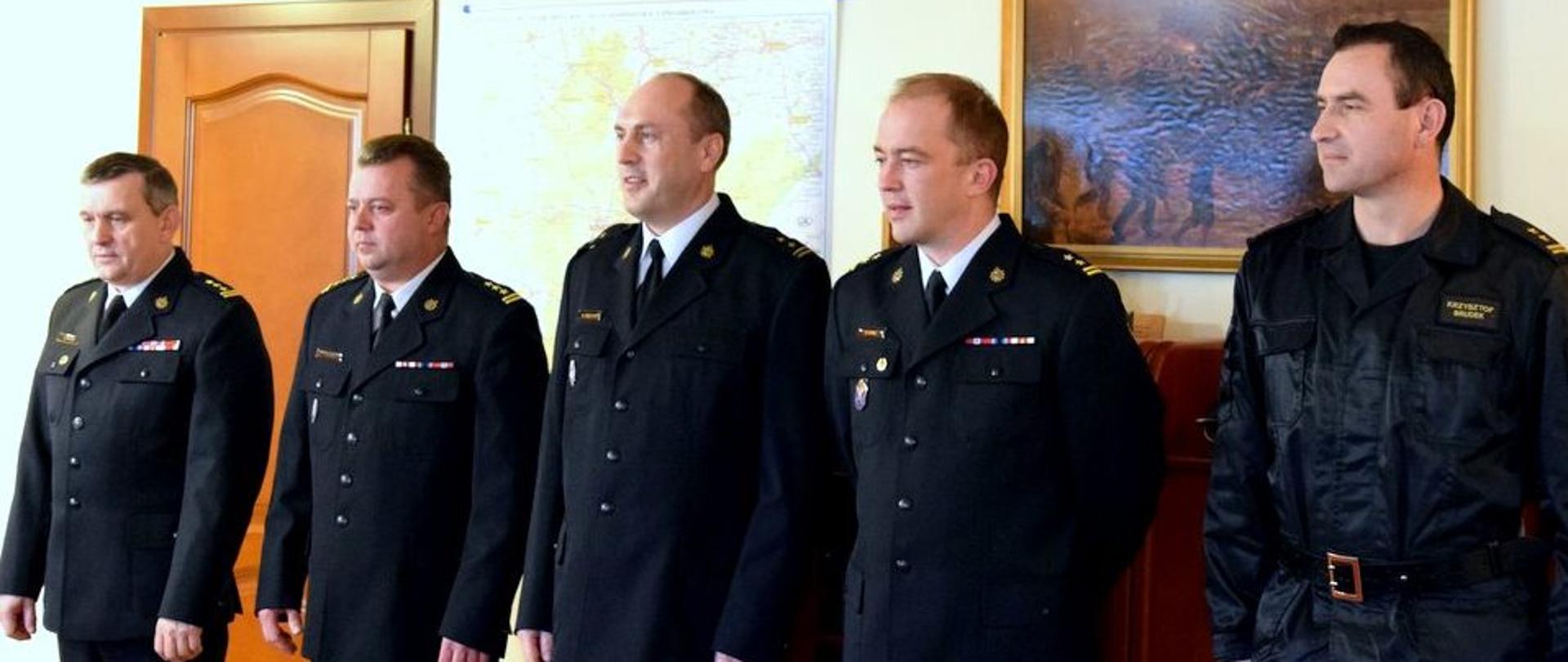 Pięciu funkcjonariuszy Państwowej Straży Pożarnej stojących w rzędzie. Strażacy są ubrani w mundury wyjściowe.
