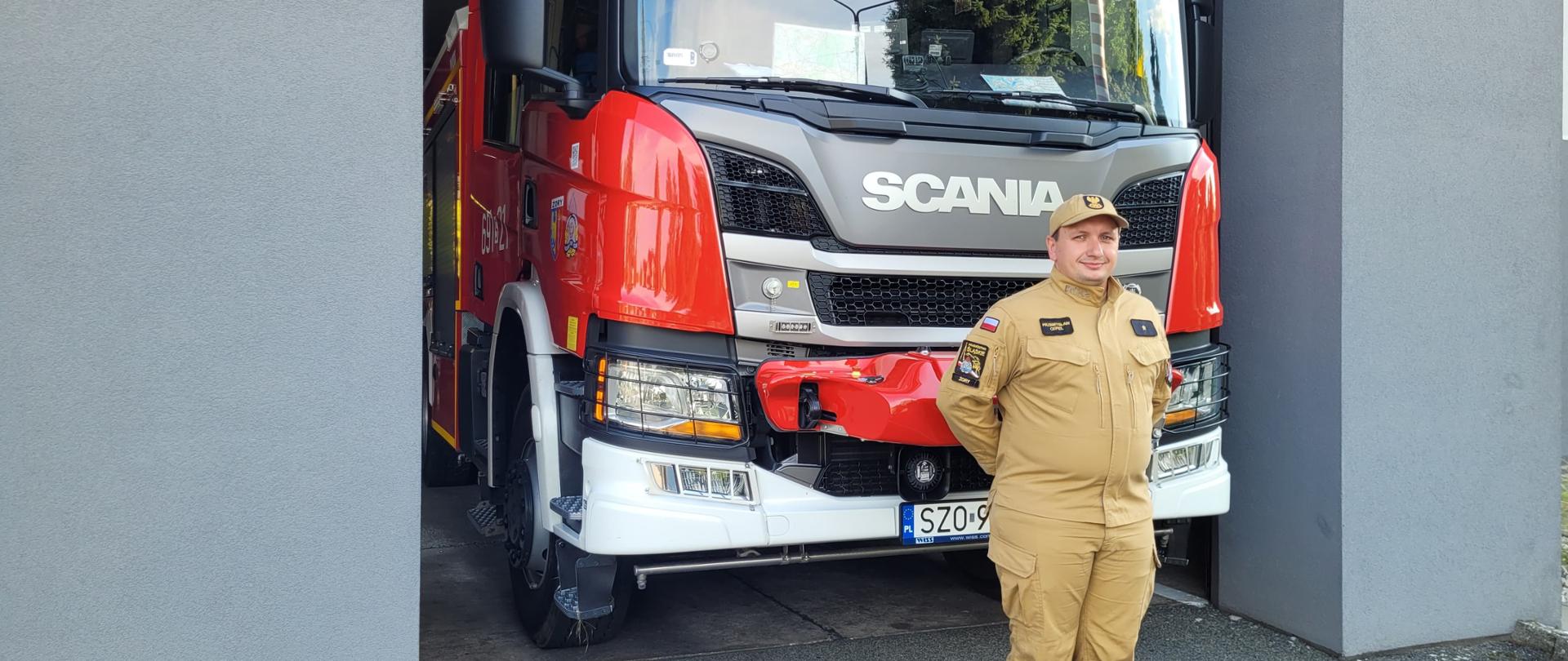 Strażak w piaskowym umundurowaniu stoi przed samochodem strażackim, który stoi w bramie garażowej. Ściany garażu są w kolorze grafitowym.