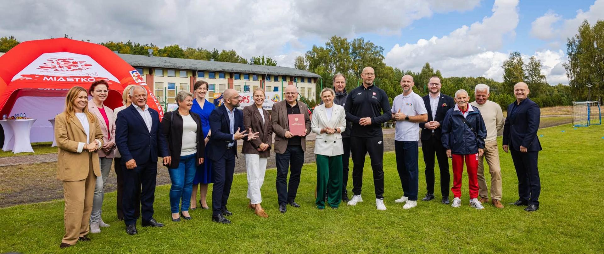 Uczestnicy konferencji w Miastku stoją na trawie. Wśród nich minister sportu i turystyki Jakub Rutnicki i ministra edukacji Barbara Nowacka. W tle budynek szkoły.