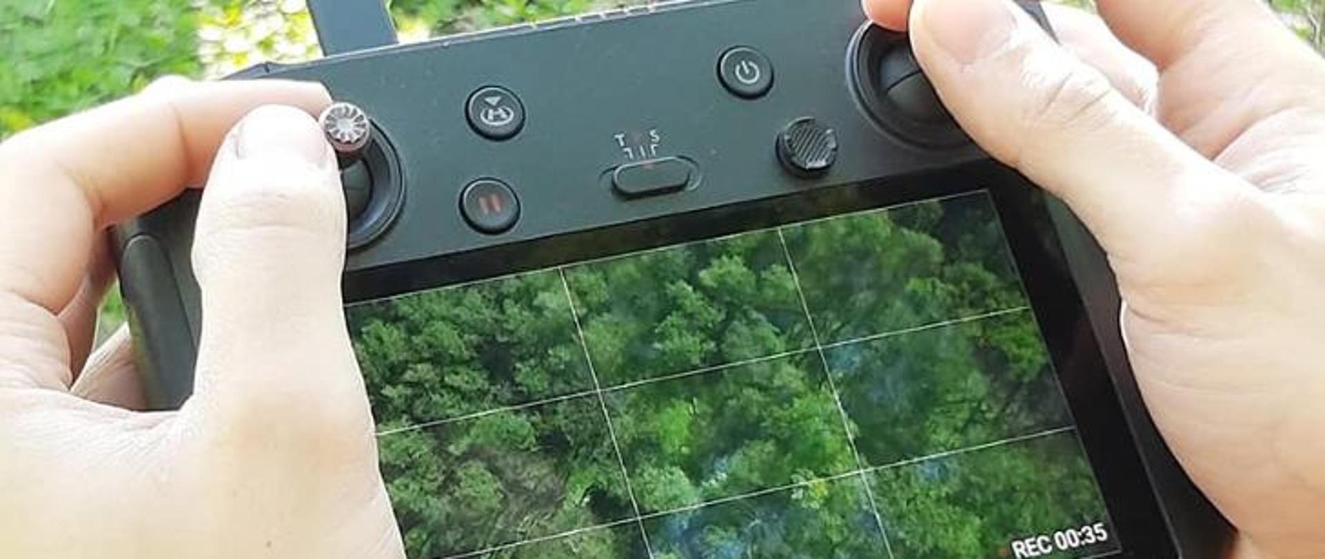 Kadr lasu z dłońmi trzymającymi prostokątne narzędzie z kamerą do obsługi drona. Urządzenie jest czarne, zawiera pokrętła i przyciski. Ma monitor, na którym widać wysokości i funkcję nagrywania REC. Tłem jest fragment lasu liściastego. Dominuje zielona kolorystyka.
