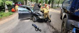 Na zdjęciu strażacy pracują przy uszkodzonym samochodzie osobowym, który zderzył się z pojazdem ciężarowym. Maska auta jest otwarta, a obok leży narzędzie hydrauliczne. W tle widoczne są służby ratownicze, osoby poszkodowane oraz zaparkowane wozy strażackie.
