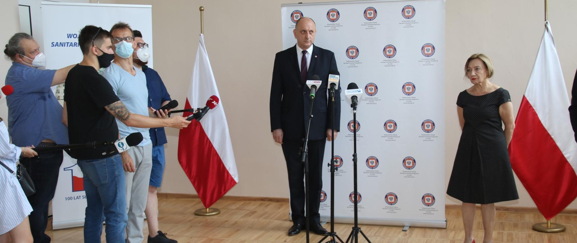 Główny Inspektor Sanitarny i Łódzki Państwowy Wojewódzki Inspektor Sanitarny stoją na tle baneru z logo Państwowej Inspekcji Sanitarnej podczas briefingu prasowego. Po lewej stronie stoi kilku mężczyzn trzymających mikrofon. Na pierwszym planie stoją trzy mikrofony. W tle zdjęcia dwie biało-czerwone flagi.