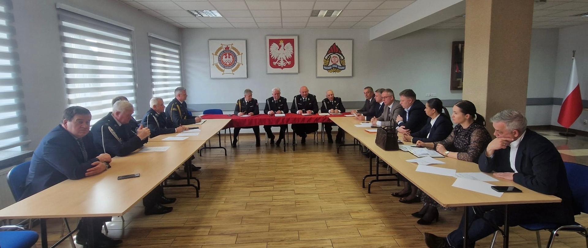 Uczestnicy posiedzenia siedzą przy ustawionych w podkowę stołach w sali konferencyjnej Komendy Powiatowej PSP w Mławie. W spotkaniu biorą udział strażacy w mundurach oraz przedstawiciele władz lokalnych, omawiając sprawy organizacyjne.