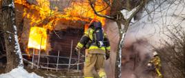 Zdjęcie przedstawia pożar drewnianego budynku mieszalnego z okna wydobywa się dym. Dwaj strażacy w ubraniach specjalnych piaskowych jeden podczas rozwinięcia linii gaśniczej.