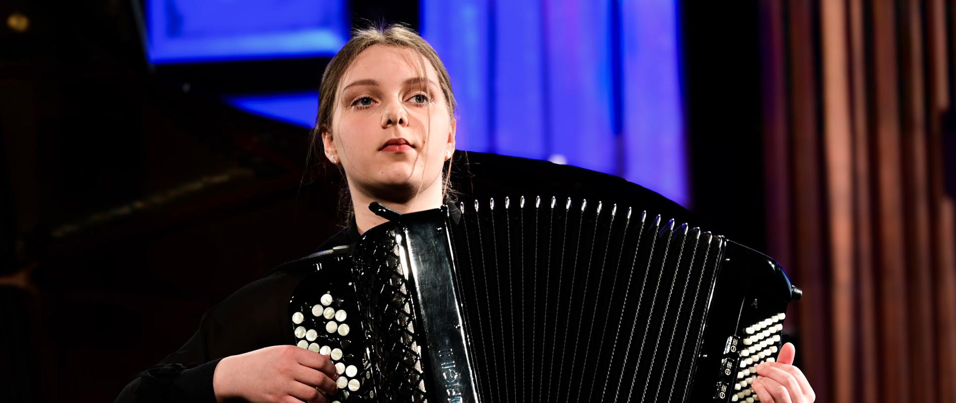Fotografia przedstawiająca młodą artystkę, Martynę Kulczycką, podczas występu na scenie. Dziewczyna siedzi i gra na dużym, czarnym akordeonie guzikowym. Jest ubrana w elegancki, czarny strój. Tło jest lekko rozmyte, z widocznymi pionowymi elementami dekoracyjnymi i niebieskim oświetleniem scenicznym, co podkreśla koncertowy charakter zdjęcia.