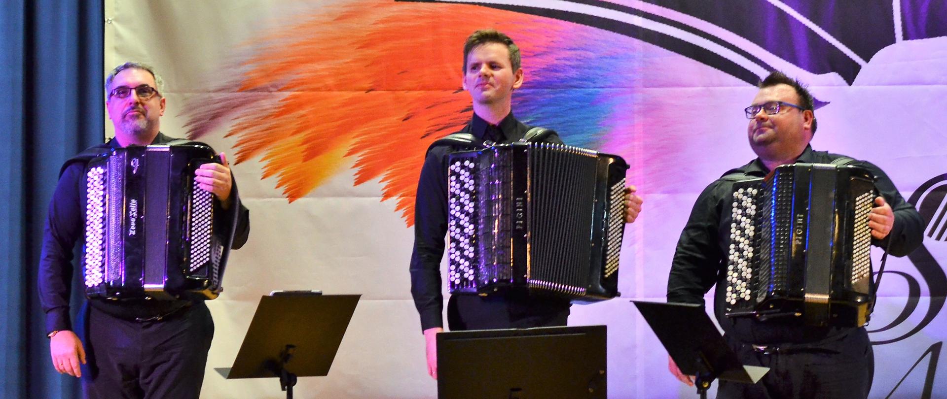 Trio akordeonowe stojące na scenie, za nimi baner konkursu akordeonowego i niebieska kotara