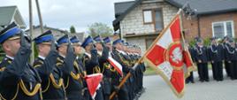Stojący w szeregu strażacy w mundurach wyjściowych salutują. Jedn z nich trzyma sztandar Komendy Powiatowej Państwowej Straży Pożarnej w Radomsku. 