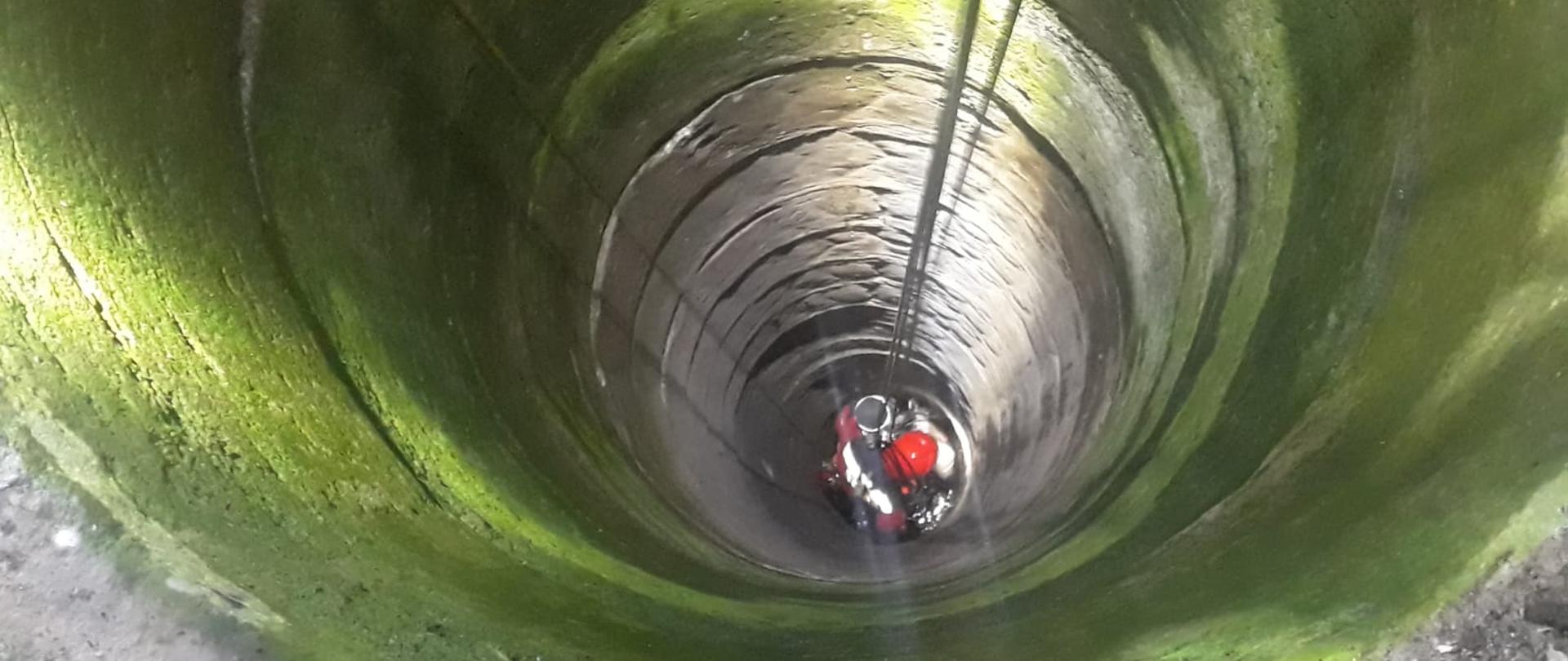 Zdjęcie przedstawia jednego z ratowników wysokościowych Specjalistycznej Grupy Ratownictwa Wysokościowego „GOŁDAP” z Komendy Powiatowej Państwowej Straży Pożarnej w Gołdapi, który zabezpieczony w aparat ochrony układu oddechowego przy wykorzystaniu technik alpinistycznych został opuszczony na dno studni w celu bezpiecznej ewakuacji uwięzionego kota na powierzchnię