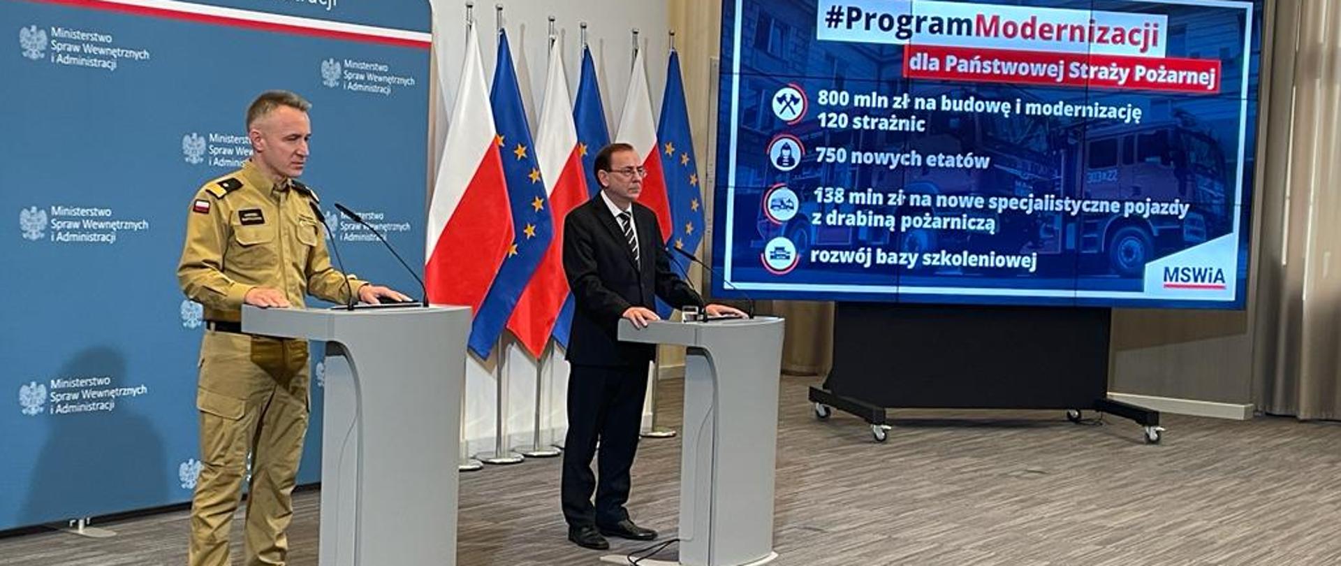 Komendant Główny PSP nadbryg. Andrzej Bartkowiak i minister Mariusz Kamiński stoją przy mównicy. Za nimi znajdują się flagi Polski oraz Unii Europejskiej.
Po lewej stronie ministra jest ukazany panel z napisem #ProgramModernizacji dla Państwowej Straży Pożarnej
