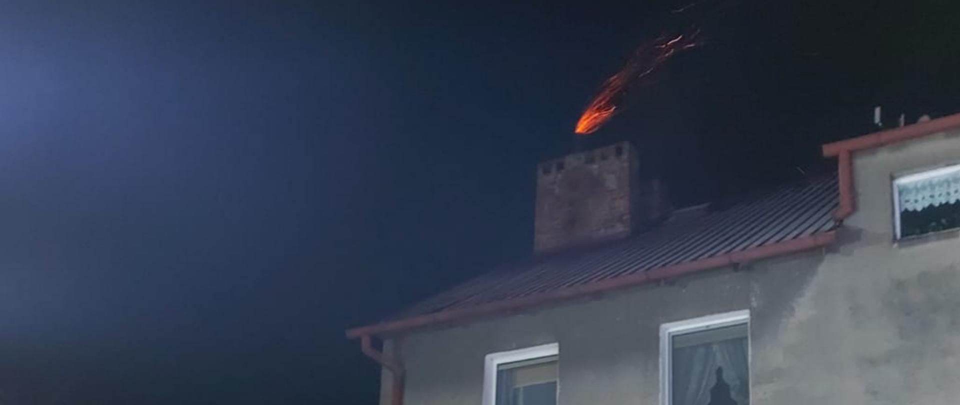 Na zdjęciu widać pożar sadzy w kominie w budynku. Z komina wydobywa się widoczny płomień w postaci czerwonej wstęgi. 