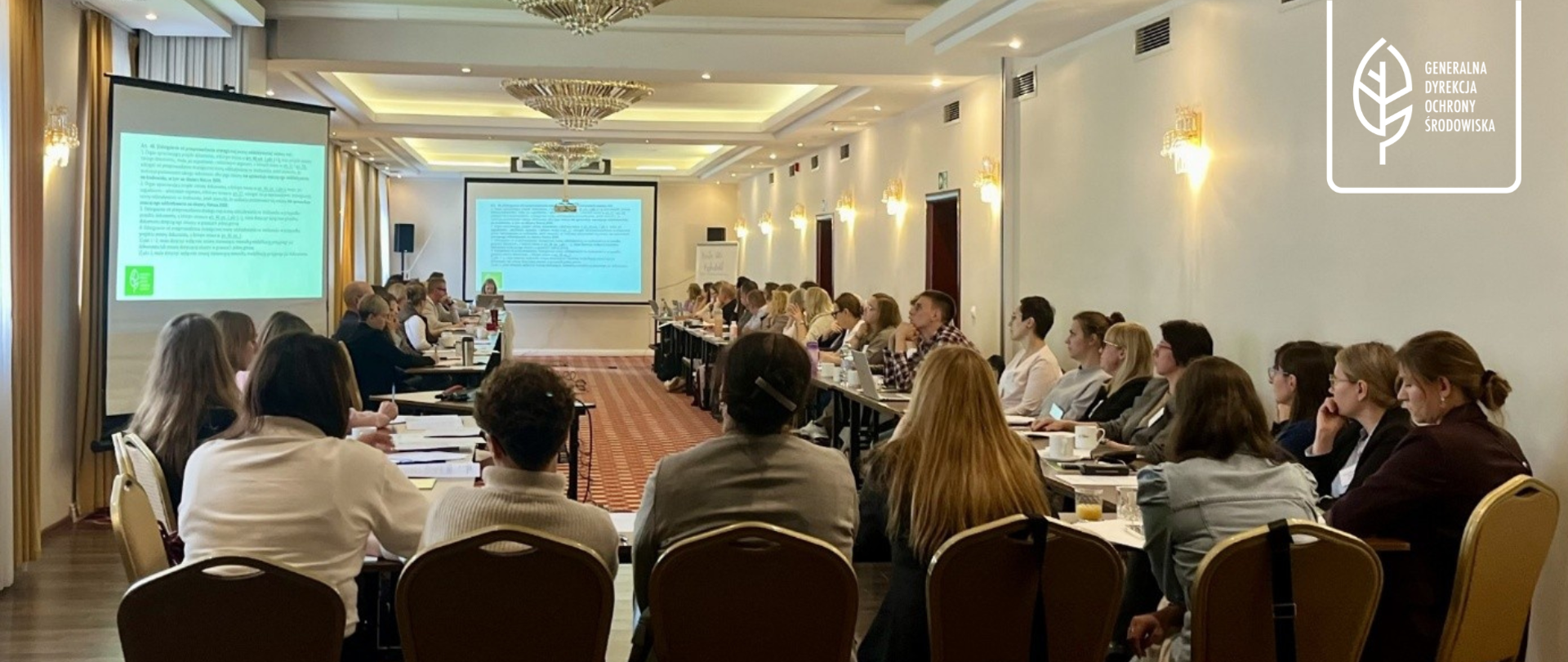 Na zdjęciu przedstawiono spotkanie warsztatowe lub szkoleniowe odbywające się w przestronnej sali konferencyjnej. Uczestnicy siedzą przy stołach ustawionych w układzie sprzyjającym dyskusji i wspólnej pracy, a przed nimi znajdują się laptopy, dokumenty i materiały szkoleniowe. W centralnej części sali wyświetlana jest prezentacja, na której skupiona jest uwaga zgromadzonych osób. Wnętrze wyróżnia się eleganckim wystrojem, jasnym oświetleniem oraz dekoracyjnymi żyrandolami. W prawym górnym rogu grafiki umieszczono logo Generalnej Dyrekcji Ochrony Środowiska.