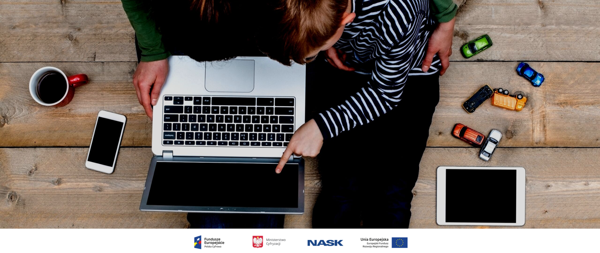 Dwie osoby siedząc patrzą się w ekran przenośnego komputera. Obok nich stoi kawa, leżą zabawki, smartfon i tablet. 