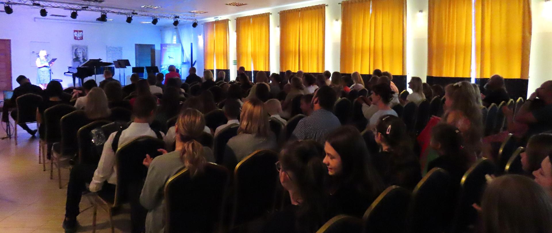 Zdjęcie przedstawia wypełnioną publicznością salę koncertową Państwowej Szkoły Muzycznej I stopnia im. J. S. Bacha w Grajewie. Po prawej stronie żółto-niebieskie zasłony, w tle scena. 