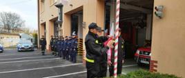 Za niskim żywopłotem 3 strażaków przypina Flagę Państwową do linki masztu. Za nimi widać budynek Jednostki ratowniczo – gaśniczej przed którą stoją w szeregu strażacy i policjanci. Widoczne są też radiowozy stojące obok budynki i częściowo samochody bojowe w budynku. 