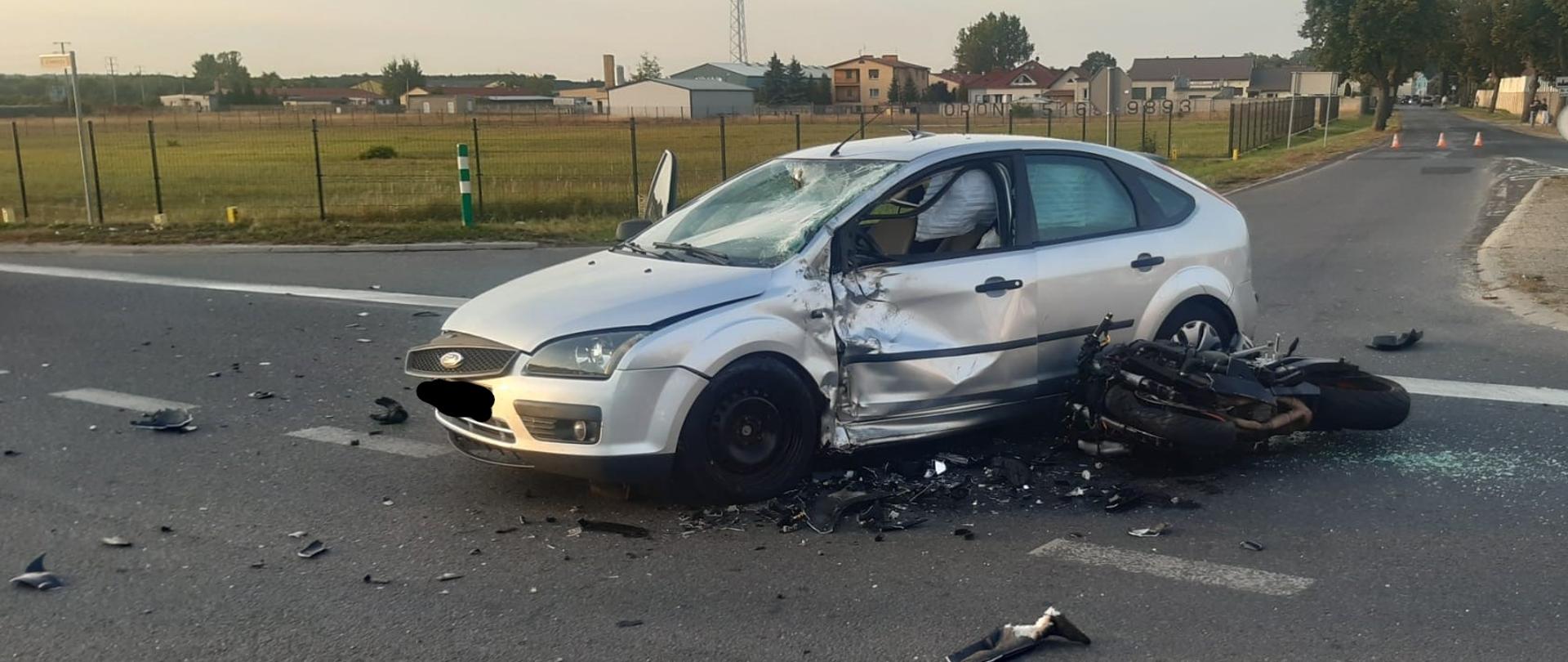 Na środku drogi stoi samochód osobowy marki ford i przy nim znajduje się uszkodzony motocykl. wokół leży rozwalone elementy motocykla.