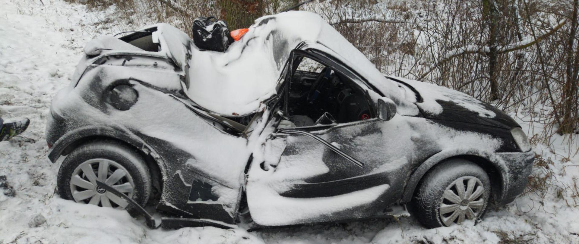 Czarny samochód marki Opel po dachowaniu, z wgiętym dachem. Znajduje się w rowie obok drogi. Pora zimowa, ziemia pokryta śniegiem. 