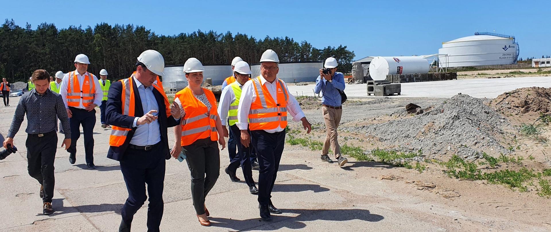 Na zdjęciu widać grupę ludzi ubranych w kaski i kamizelki odblaskowe, Ci ludzie są na budowie. Pogoda jest słoneczna, niebo niebieskie - bez chmur.