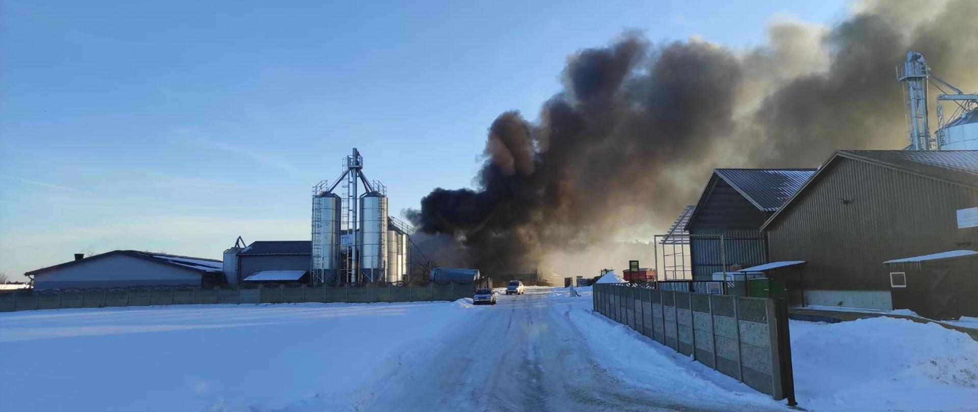 Zdjęcie zrobione w trakcie dojazdu do pożaru, widać kłęby dymy wydostające się z budynku gospodarczego z bezpośrednim sąsiedztwie z budynkami inwentarskimi. Widać duże silosy zbożowe przy budynku inwentarskim droga do pożaru utrudniona ,oblodzona powierzchnia drogi. Po drugiej stronie pożaru stoją wiaty metalowe które nie są zagrożone pożarem.
