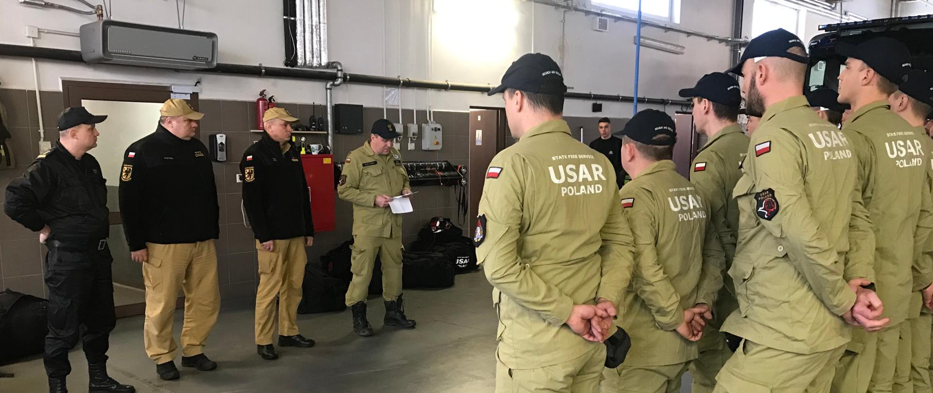 Strażacy w garażu jednostki ratowniczo-gaśniczej w strojach z napisem USAR Poland stoją w rzędzie przed kierownictwem Komendy Wojewódzkiej Państwowej Straży Pożarnej w Gdańsku. 