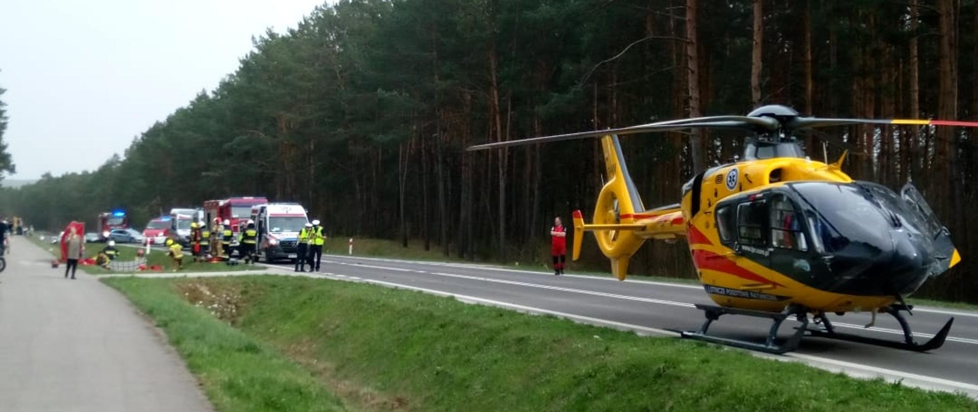 Zdjęcie przedstawia śmigłowiec stojący na pasie drogowym, a w głębi widać pojazdy służb ratowniczych oraz ratowników podczas działań.