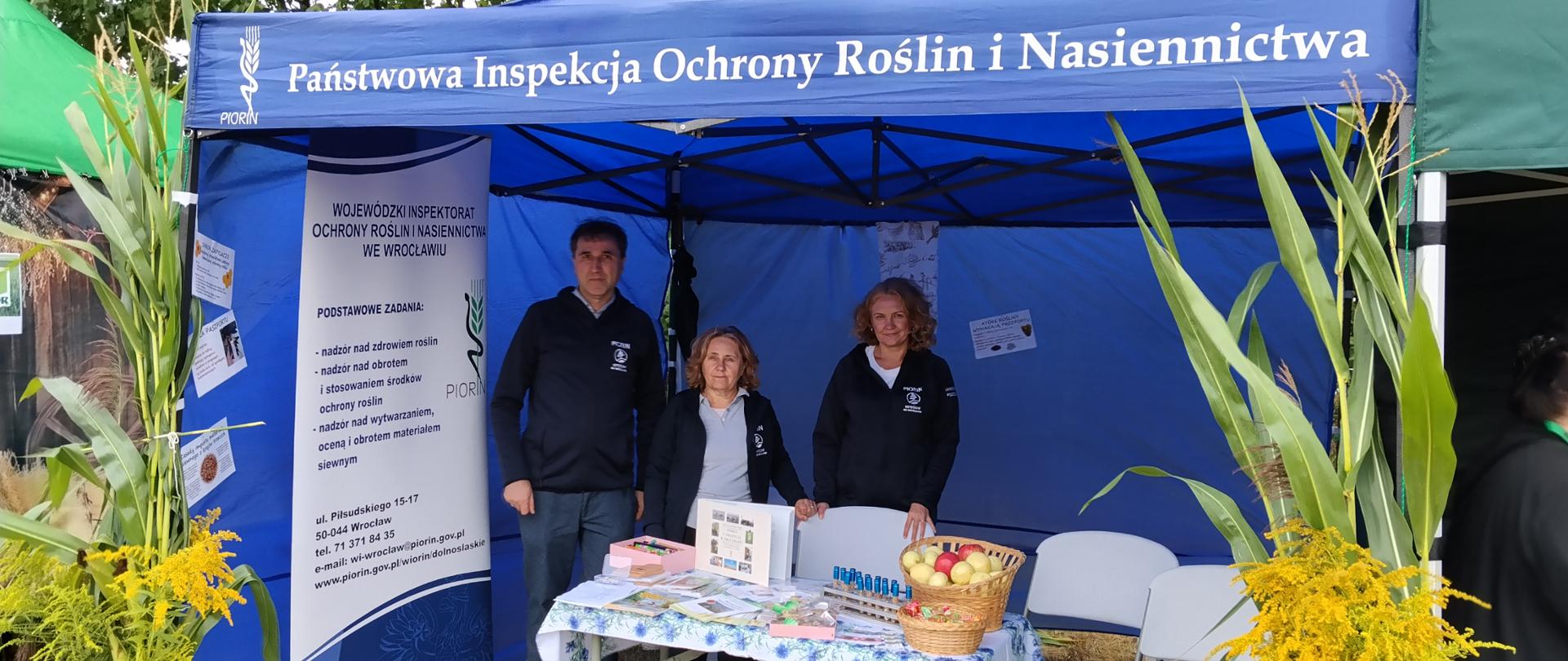 Stoisko Państwowej Inspekcji Ochrony Roślin i Nasiennictwa pod niebieskim namiotem. Przed stoiskiem stoi troje inspektorów (dwie kobiety jeden mężczyzna) a na stole znajdują się materiały informacyjne, produkty oraz kosz z owocami. Stoisko otaczają wysokie rośliny i żółte kwiaty.