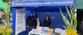 Stoisko Państwowej Inspekcji Ochrony Roślin i Nasiennictwa pod niebieskim namiotem. Przed stoiskiem stoi troje inspektorów (dwie kobiety jeden mężczyzna) a na stole znajdują się materiały informacyjne, produkty oraz kosz z owocami. Stoisko otaczają wysokie rośliny i żółte kwiaty.