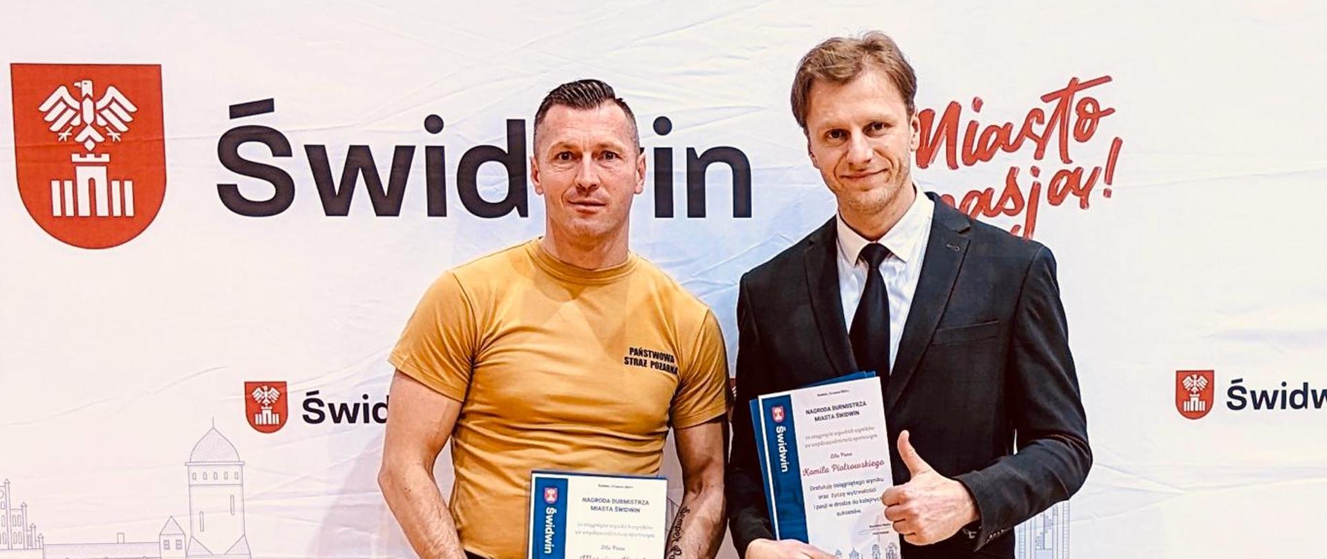 Zdjęcie przedstawia strażak podczas wręczenia nagrody, pozuje z mężczyzna na tle scianki