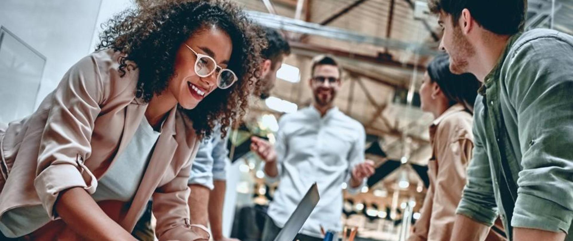 Kobieta pisząca na komputerze, pozostałe 4 osoby zgromadzone wokół stołu 