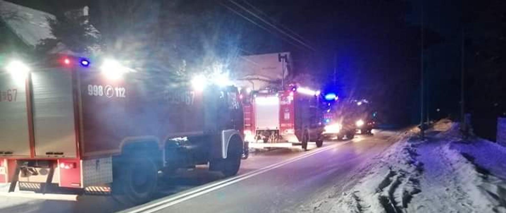 Zdjęcie przedstawia cztery zastępy jednostek ochrony przeciwpożarowej, ustawionej na drodze asfaltowej jeden za drugim w zimowej scenerii z włączoną sygnalizacją świetlną błyskową o barwie niebieskiej. 