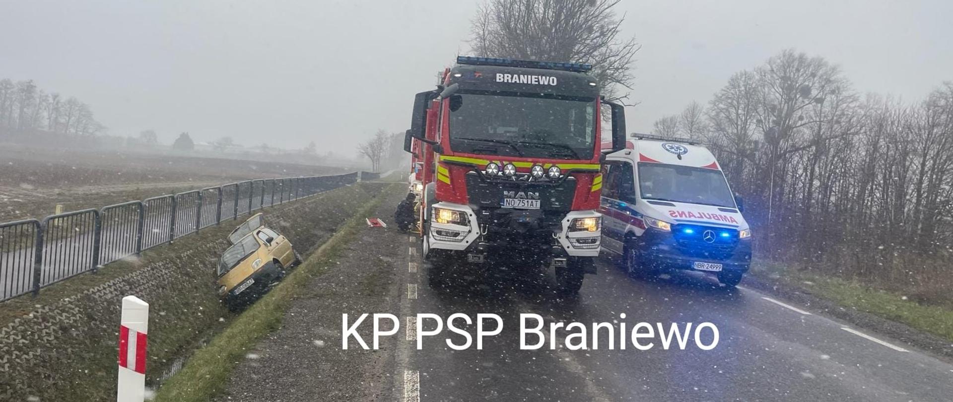 Pochmurno, pada śnieg. Z lewej strony na zdjęciu auto osobowe w rowie. Na środku stoi samochód strażacki, obok karetka pogotowia.