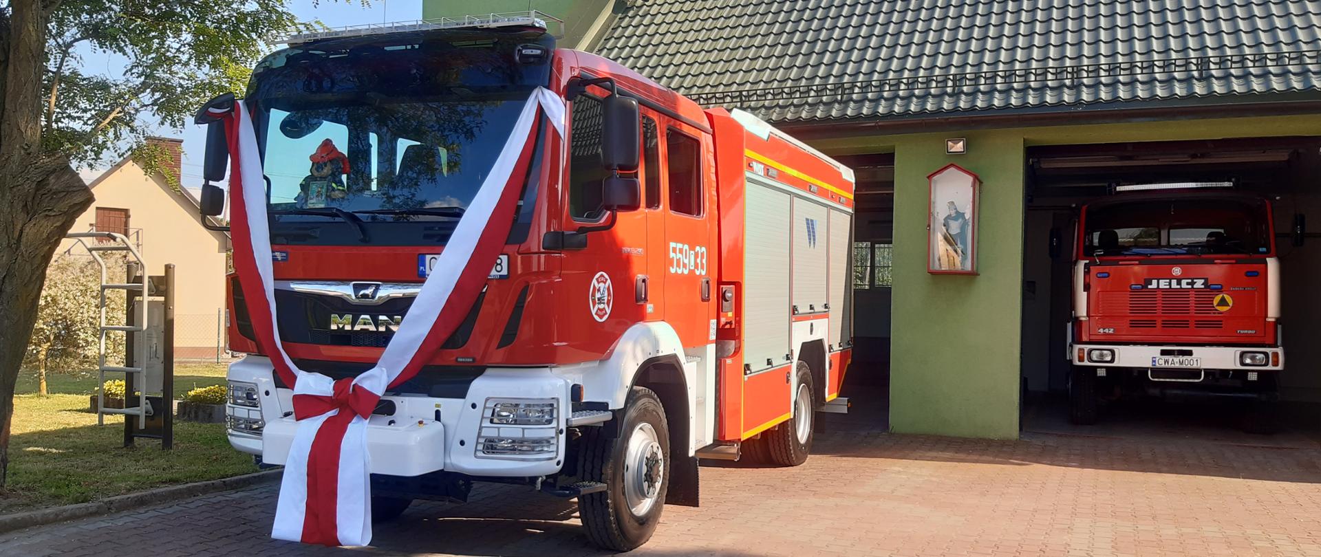 Na pierwszym planie wóz strażacki, udekorowany wstęgą biało-czerwoną. W tle obiekt remizy.