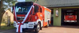 Na pierwszym planie wóz strażacki, udekorowany wstęgą biało-czerwoną. W tle obiekt remizy.
