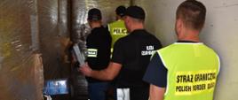 Funkcjonariusze stoją w naczepie pojazdu ciężarowego i oglądają kartony z papierosami.