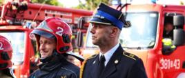 Strażacy w mundurach bojowych w kolorze granatowym i piaskowym oraz strażak w mundurze galowym stoją na placu JRG 2. W tle czerwone wozy strażackie 