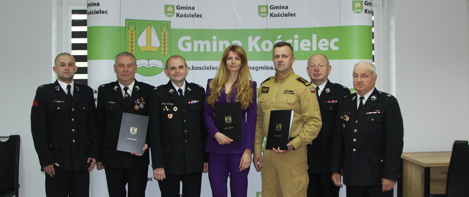 Zdjęcie przedstawia siedem osób, stojących przed zielono-białym banerem z logo Gminy Kościelec. W grupie jest pięciu mężczyzn w mundurach służb ratowniczych, kobieta w fioletowym garniturze oraz jeden mężczyzna w beżowym mundurze. Niektórzy z nich trzymają teczki lub dokumenty. Wszystkie osoby patrzą w stronę obiektywu, pozując do zdjęcia.