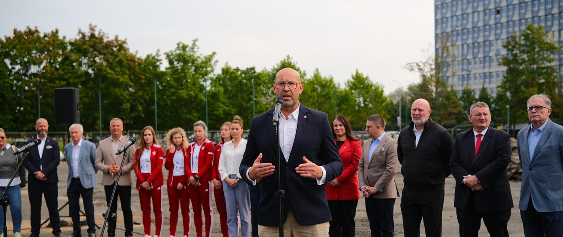Minister Jakub Rutnicki na terenie Politechniki Poznańskiej. Stoi za mikrofonem na tle budynku politechniki. Za nim stoją włodarze miasta, trenerzy i zawodnicy
