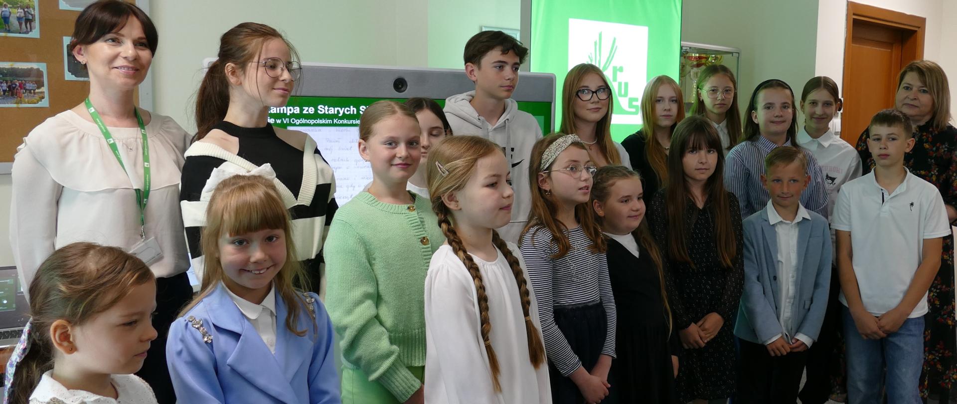 W sali oddziału KRUS grupa dzieci pozuje do zdjęcia z dwiema kobietami. Wszyscy uśmiechają się do zdjęcia. Z tyłu za nimi monitor i ekran z zielonym logo KRUS.