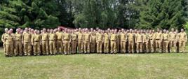 Zdjęcie grupowe kilkudziesięciu strażaków mundurach piaskowych stojących na trawniku przed wysokimi zielonymi drzewami. 