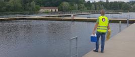 Osoba stojąca na betonowym pomoście kąpieliska. Osoba ubrana w żółtą kamizelkę z logo Państwowej Inspekcji Sanitarnej trzyma w rękach sprzęt do poboru próbek wody.