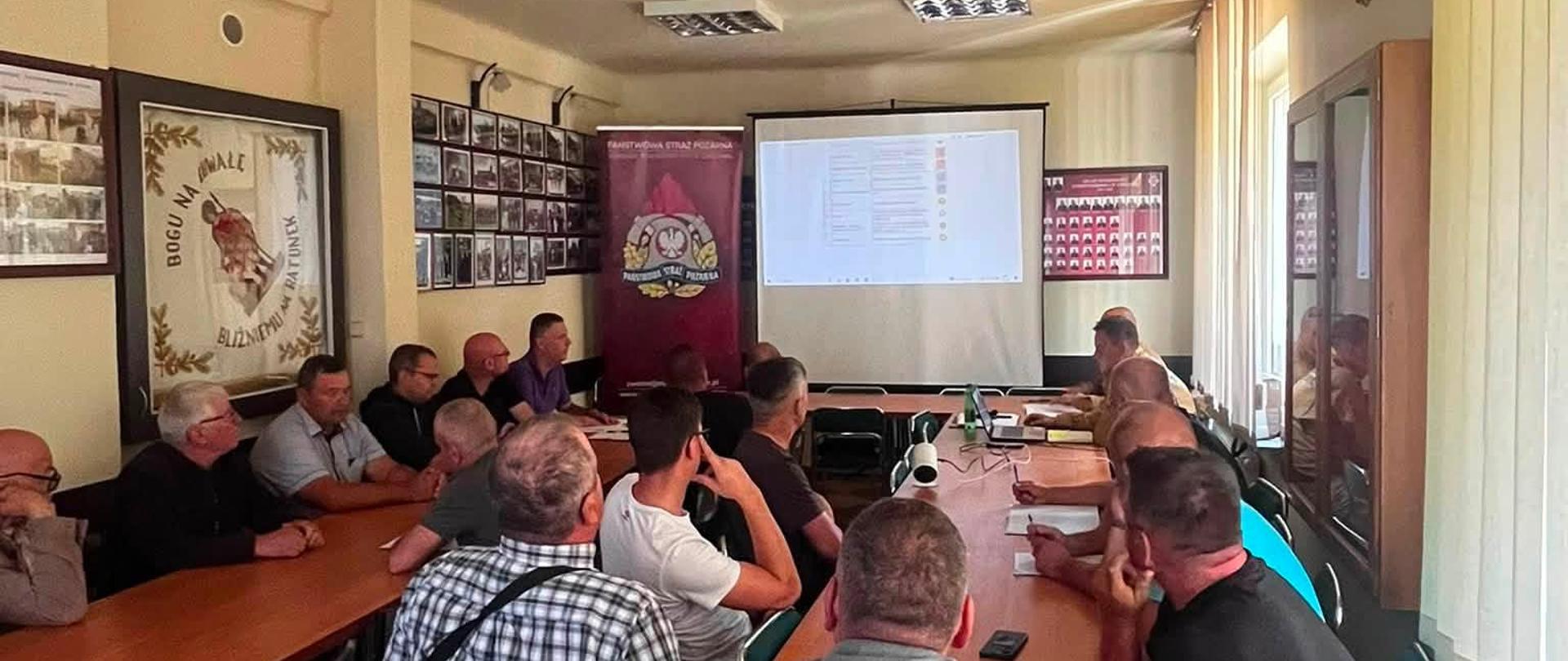 Fotografia wnętrza sali szkoleniowej lub konferencyjnej, w której odbywa się spotkanie. Wzdłuż długiego, drewnianego stołu siedzi kilkunastu mężczyzn, w większości ubranych w stroje cywilne (koszule, t-shirty), skierowanych twarzami w stronę ekranu projekcyjnego na końcu sali. Niektórzy robią notatki. Na ekranie wyświetlana jest prezentacja lub dokument. Obok ekranu stoi pionowy banner (roll-up) z logo Państwowej Straży Pożarnej. Po lewej stronie, na ścianie, wisi duży, oprawiony proporzec z wyhaftowanym orłem i napisem: "BOGU NA CHWAŁĘ, BLIŹNIEMU NA RATUNEK". Ściana powyżej udekorowana jest licznymi, mniejszymi, oprawionymi zdjęciami. Sala jest oświetlona podłużnymi lampami sufitowymi oraz światłem dziennym wpadającym przez okno po prawej.