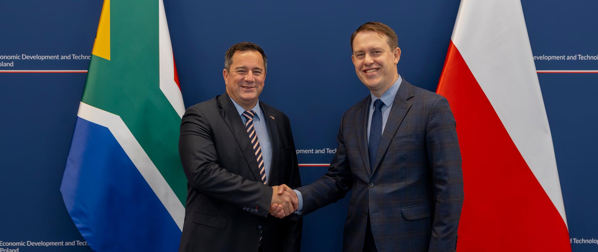 Od lewej strony stoją na tle flag RPA i RP wiceminister handlu, przemysłu i konkurencji RPA, John Steenhuisen oraz wiceminister rozwoju i technologii, Michał Baranowski; W tle ścianka ministerstwa handlu przemysłu i technologii; politycy podają sobie ręce