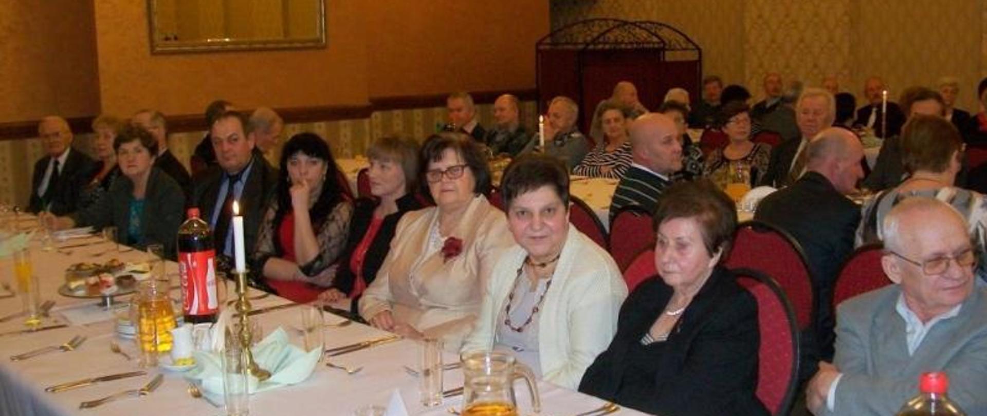 Na zdjęciu widzimy kilkadziesiąt osób mężczyzn i kobiet siedzących przy stole. Stół nakryty jest białym obrusem we wzory, na stole widzimy talerze, szklanki, napoje oraz serwetki. Osoby znajdują się w pomieszczeniu, dalej widzimy wystrój tego wnętrza, ściany pokryte żółtą tapetą we wzorki. Na ścianie widzimy też świecące kinkiety oraz lustro w złotej oprawie. Z sufitu zwisają brylantowe żyrandole.