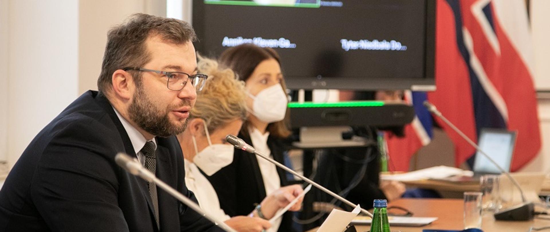 Minister Grzegorz Puda siedzi przy stole konferencyjnym i mówi do mikrofonu. Obok siedzą dwie kobiety, w dalszym planie widoczny ekran.