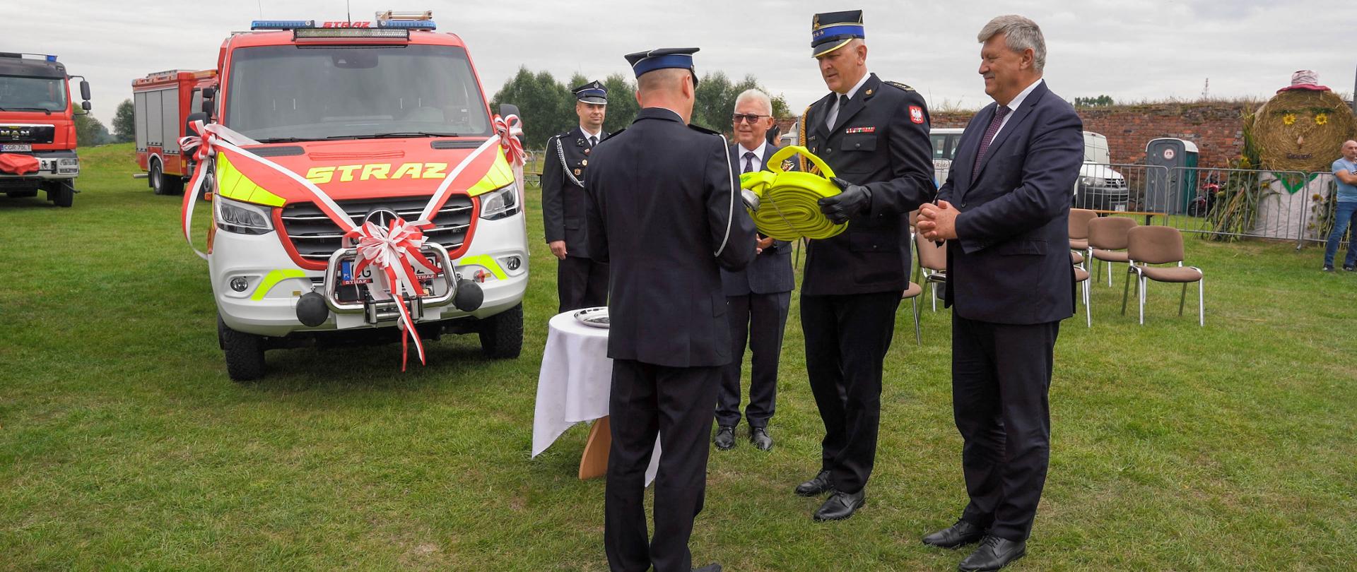 Czerwony pojazd strażacki zaparkowany na trawiastym terenie. Obok niego stoją strażacy i osoby cywilne. jeden strażak wręcza drugiemu strażakowi żółty wąż. W tle inne pojazdy.