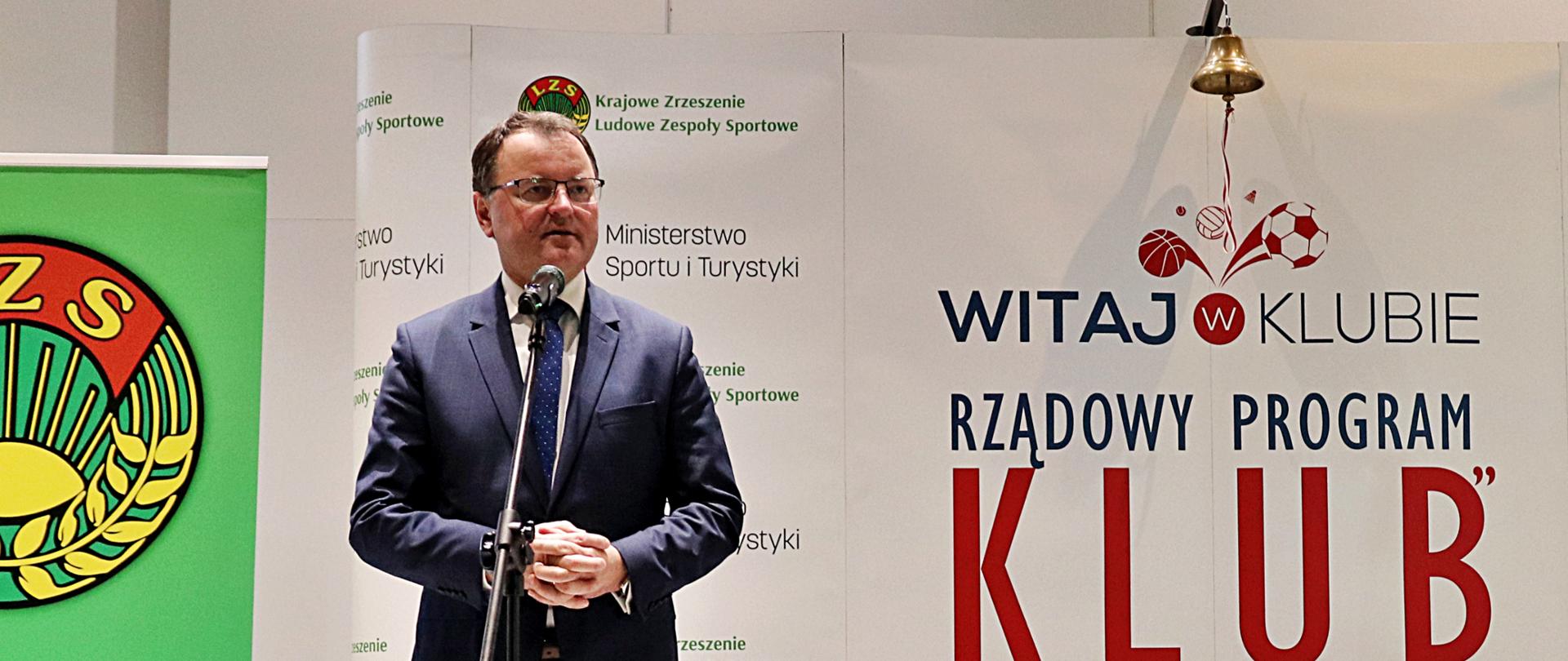 Na zdjęciu znajduje się elegancko ubrany mężczyzna. Mężczyzna stoi przed mikrofonem i jest w trakcie wystąpienia publicznego. W tle znajdują się materiały wizerunkowe Rządowego Programu KLUB.