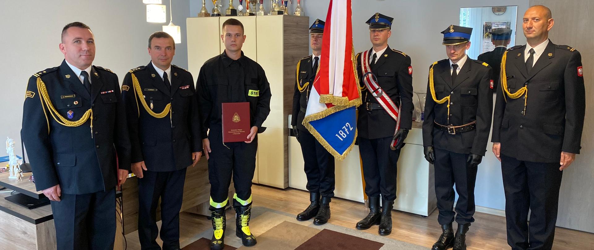 Zdjęcie przedstawia nowo przyjętego strażaka z reprezentacją Komendy Miejskiej Państwowej Straży Pożarnej w Rzeszowie, komendantem miejskim Państwowej Straży Pożarnej w Rzeszowie oraz pocztem sztandarowym w gabinecie komendanta miejskiego PSP w Rzeszowie. Wszystkie osoby przyjęły w postawę zasadniczą. 