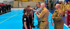 Podczas II Halowych Młodzieżowych Zawodów Sportowo-Pożarniczych przedstawiciel PSP gratuluje młodym zawodnikom stojącym w szeregu na hali sportowej.

