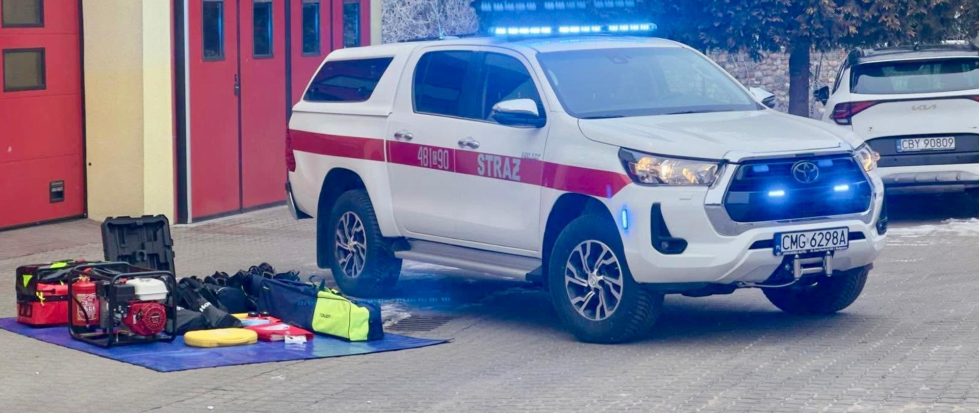 Nowy samochód w KP PSP Mogilno. Po lewej stronie na płachcie leżą pozyskane z samochodem sprzęty. Po prawej stronie nowy samochód z włączonymi światłami 